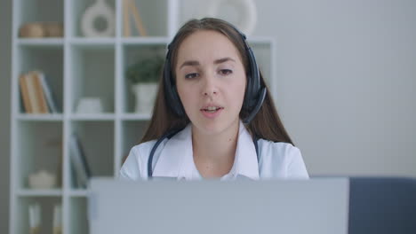 Female-medical-assistant-wears-white-coat-headset-video-calling-distant-patient-on-laptop.-Doctor-talking-to-client-using-virtual-chat-computer-app.-Telemedicine-remote-healthcare-services-concept.